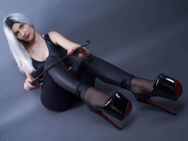 MistressKate with silver hair, black leather top, tight black pants, sheer black stockings, and black high heels with red soles, holding a black riding crop, sitting on the floor.