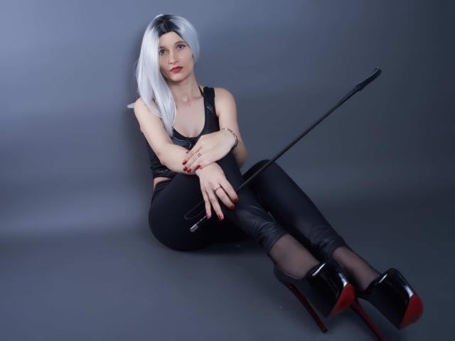 MistressKate with white hair, black leather top, black leather pants, black high heels, red nails, holding a black whip, sitting on gray floor, legs crossed, looking at camera.