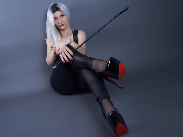 MistressKate with white hair, black leather outfit, and red-soled heels, holding a black whip, sitting with legs crossed, showing her ass and black boots with red soles.