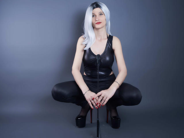 MistressKate with white hair, black leather zip-up top, black pants, and high heels, squatting, holding a black whip, red nails, and a confident smile, showing her tits.