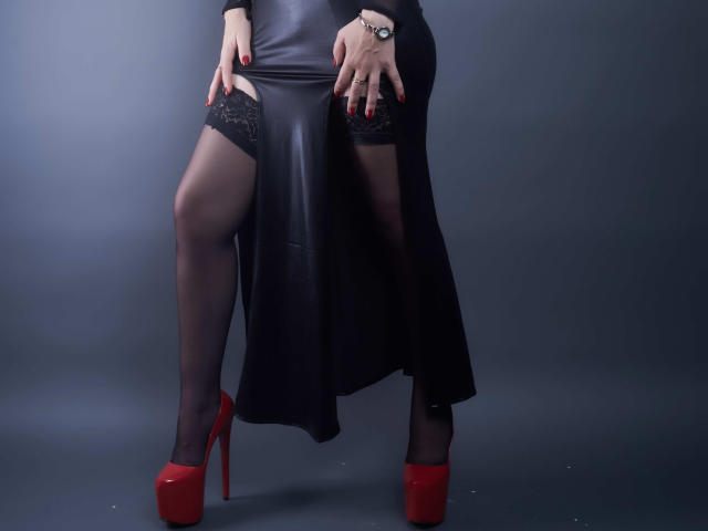 La robe en cuir noir de MistressKate, ses bas en dentelle noire et ses talons rouges mettent en valeur ses jambes, et elle pose ses mains sur ses cuisses, tout en portant une montre noire..