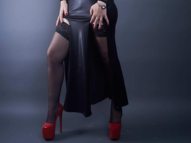 La jupe en cuir noir de MistressKate, ses bas noirs transparents et ses hauts en dentelle rouge, avec ses talons rouges, elle se tient debout, les mains sur ses cuisses, montrant ses ongles peints en rouge..