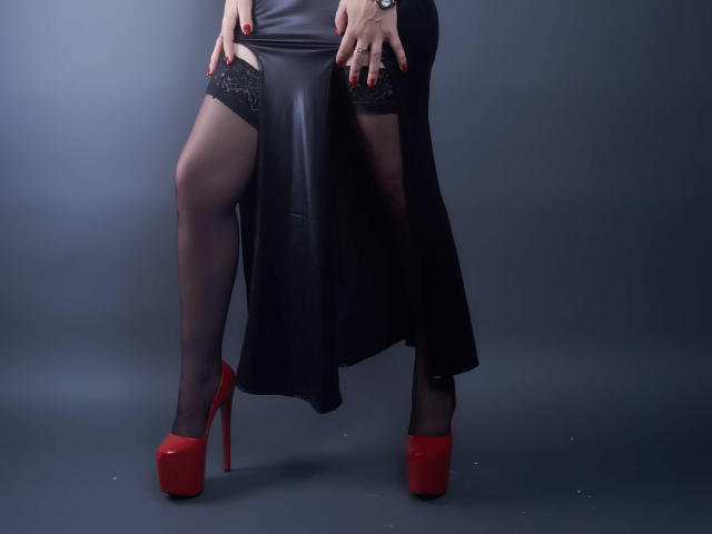 Les jambes de MistressKate, avec des bas en dentelle noire, des talons rouges et une robe en cuir noir, mettant en valeur ses cuisses et ses mains avec des ongles rouges. Fond noir..