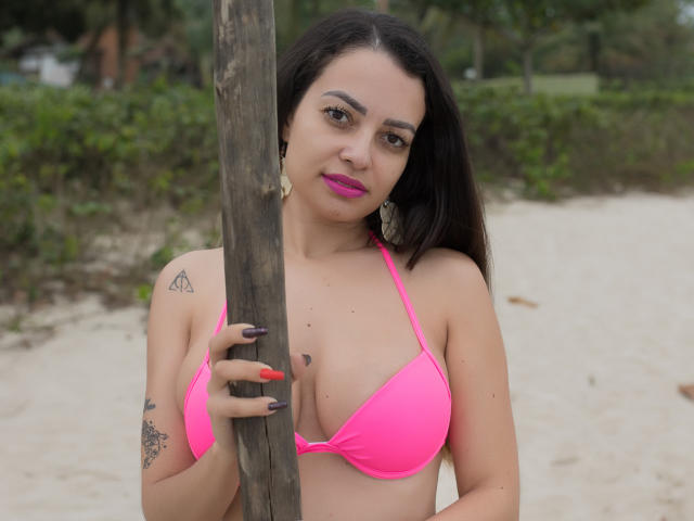 Amara, con un bikini rosa, sosteniendo un palo de madera, tiene tatuajes, lápiz labial rosa, cabello negro largo y uñas rosas, está de pie en una playa arenosa, mirando a la cámara..