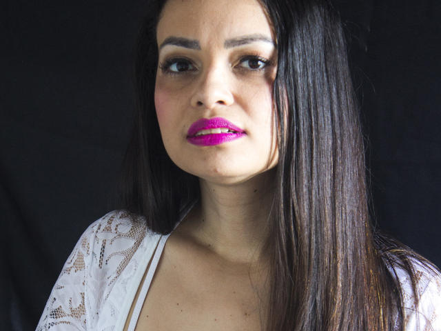 AmaraLove has dark straight hair, pink lipstick, and a white lace top with a low neckline. She's staring with a slightly concerned expression.