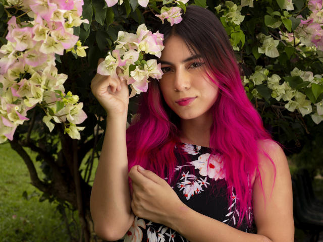 JuliaGarciax with bright pink hair, wearing a black floral dress, holding pink and white flowers, smiling, pink lipstick, green eyes, standing in front of a blooming bush.