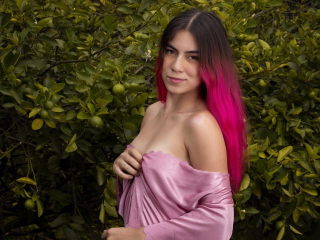 JuliaGarciax with pink-to-brown gradient hair, off-shoulder pink dress, holding dress down to expose small tits, smiling at camera, standing in front of green leafy background.