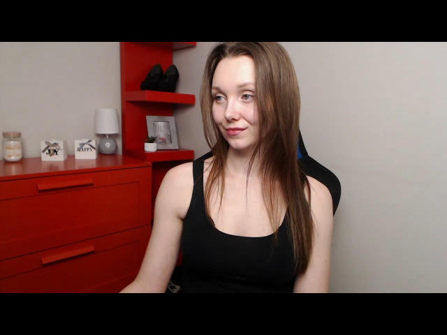 QueenZoe, avec de longs cheveux bruns, de petites seins, portant un débardeur noir, est assise dans une pièce avec des tiroirs et des étagères rouges. Elle sourit légèrement et regarde l'appareil photo..