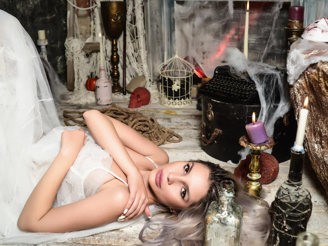 GoddessKaterina lying on her back with white dress, hands on chest, surrounded by candles, a black typewriter, and smoke. Her tits are visible, she has blonde hair.