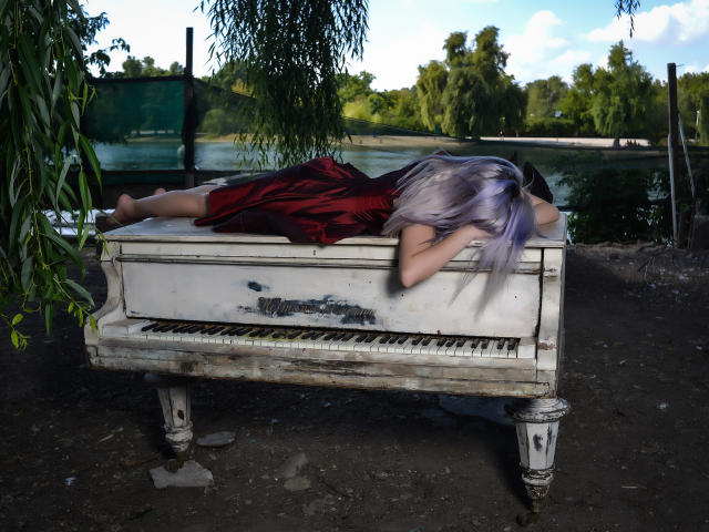 GoddessKaterina with purple hair, lying face-down on a dirty, white, old piano, wearing a red dress, her ass up, bare feet, outside in a grassy, tree-filled area.