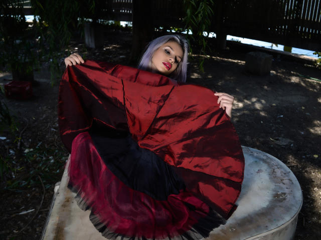 GoddessKaterina with purple hair, red and black dress, holding up the red part, sitting on a circular stone, in a dimly lit, wooded area. Her tits are covered.