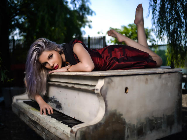 GoddessKaterina with silver hair, lying on a dirty white piano, wearing a red satin dress, legs up, looking seductively at the camera, bare feet, gold bracelet.