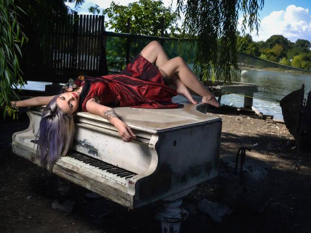 GoddessKaterina with purple hair, lying on an old piano, wearing a red dress, black heels, and black necklace, her tits slightly visible, by a sunny lake.