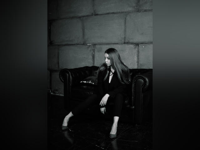 PolinaJonesss is a sexy woman with long hair, sitting on a black leather couch, wearing a black outfit and flats, looking to the side in a black-and-white photo.
