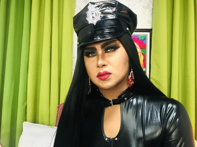 BornStarTS in a shiny black leather outfit, wearing a police cap, with long black hair, green eyes, and red lipstick, looking serious. Her tits are small, and she's got red earrings.