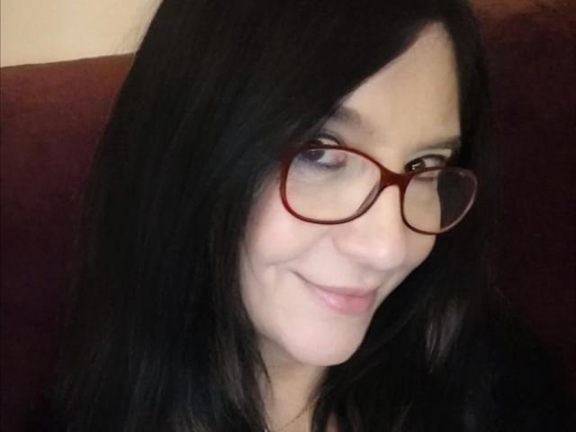 Emilianne tem cabelo preto, óculos vermelhos e um leve sorriso, com pele clara e maquiagem discreta. Seus olhos estão voltados para o lado, e ela está vestindo uma blusa escura..