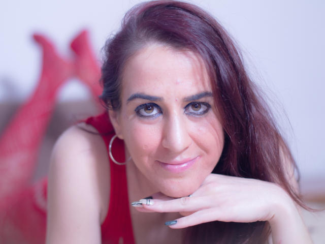 KynkyPeach hat rotes Haar, braune Augen und ein leichtes Lächeln, liegt auf der Brust, wobei ihre Hand unter ihrem Kinn ruht, und trägt rote Spitze und rote Netzstrümpfe..