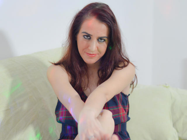 KynkyPeach, con cabello rojo, vestida con un vestido de cuadros, está sentada en un sofá verde, con los brazos cruzados, sonriendo ligeramente, con los ojos mirando hacia adelante, y con reflejos de luz en su piel..