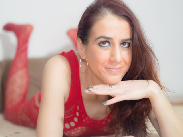 KynkyPeach con medias de red rojas, acostada en una cama, sonriendo, con la mano debajo del mentón, vistiendo una blusa de encaje roja, aros grandes y uñas verdes..