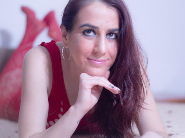 KynkyPeach met lang bruin haar, een rode jurk, oorringen en een subtiele glimlach, leunt met haar kin op haar handen, toont haar borsten en kijkt recht in de camera..