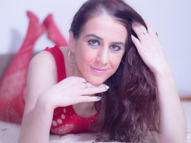 KynkyPeach, met bruin haar en rode lingerie, leunt tegen een bed en glimlacht naar de camera. Ze draagt oorringen, ringen en heeft een hand met gelakte nagels. Haar borsten zijn zichtbaar..