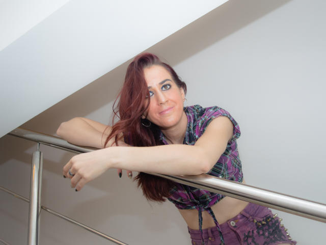 KynkyPeach, avec ses cheveux roux, se penche contre une rambarde en métal, vêtue d'une chemise à carreaux et d'un pantalon violet, et sourit avec ses yeux bleus. Ses seins sont visibles sous la chemise..