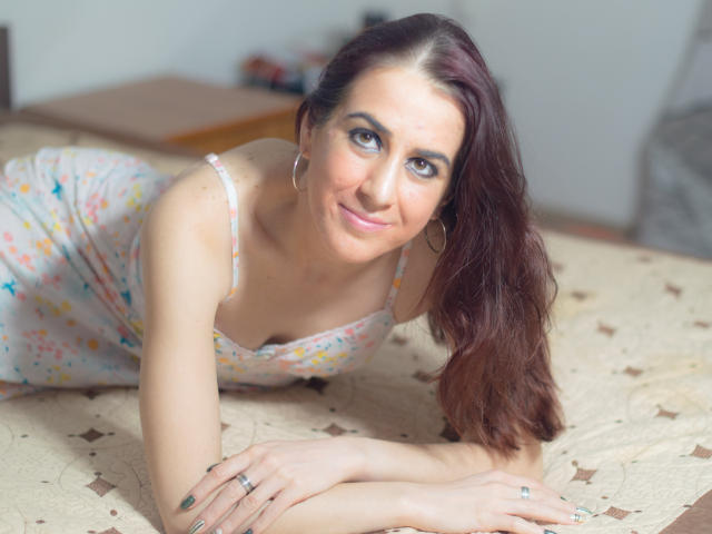 KynkyPeach, con el pelo largo y castaño, está acostada en una cama, vestida con un vestido floral, sonriendo a la cámara, con aros grandes y un anillo en su mano izquierda..