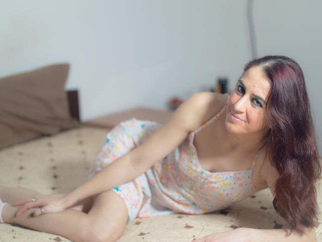 KynkyPeach est allongée sur un lit, vêtue d'une robe à fleurs, montrant ses seins, avec de longs cheveux bruns et un léger sourire, dans une pièce éclairée doucement..