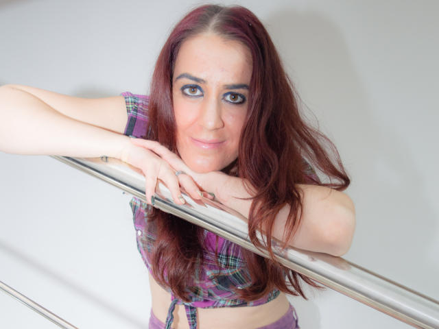 KynkyPeach, con cabello rojo, una blusa a cuadros y ropa interior morada, apoyada en una barandilla de metal, sonriendo, mirando a la cámara, mostrando pequeños senos, piel clara y maquillaje azul en los ojos..