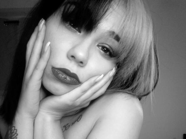 KassiKassou hat dunkel- und hellblondes Haar, viel Make-up, glänzende Lippen und Tattoos an ihren Händen. Sie berührt ihr Gesicht mit lackierten Nägeln, auf einem Schwarz-Weiß-Selfie..