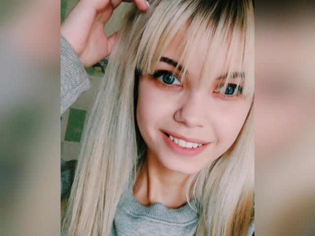 MiraCandy with blonde hair, blue eyes, and a cute smile, wearing a grey sweater, touching her head, showing off her small nose and white teeth.