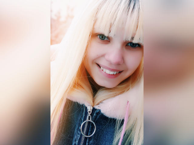 MiraCandy, com cabelo loiro, olhos azuis e um sorriso adorável, vestindo uma jaqueta preta com zíper e detalhes de pele rosa, e um zíper prateado, olhando diretamente para a câmera..