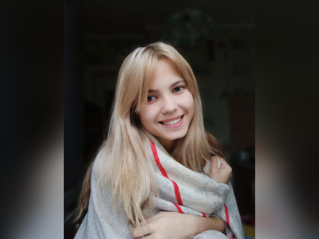 MiraCandy, com cabelo loiro, sorrindo, vestindo um cobertor cinza com listras vermelhas, cobrindo seus seios, em pé em um quarto escuro, com uma luz suave em seu rosto..