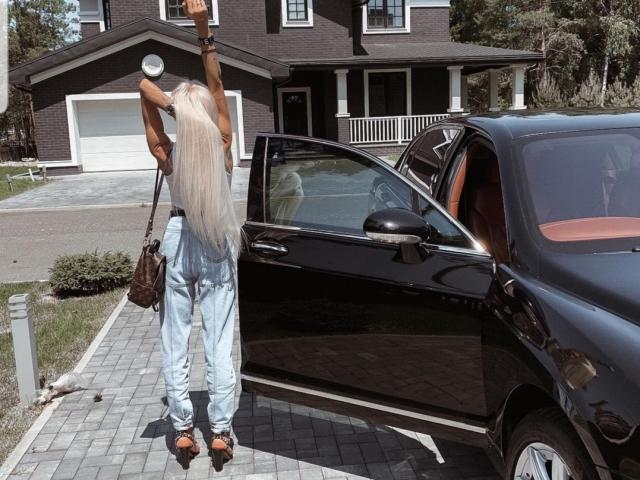 LilianaWax with long blonde hair, arms raised, wearing light blue pants and brown sandals, standing beside a black car with door open, holding a brown bag.