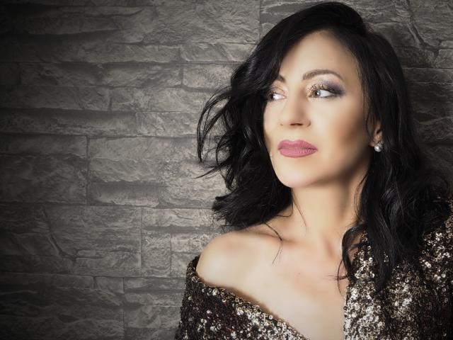 MistressTaylor with wavy black hair, sparkly makeup, pink lipstick, pearl earrings, and sequined brown top, looking to the side, showing off her tits and elegant, sexy face.