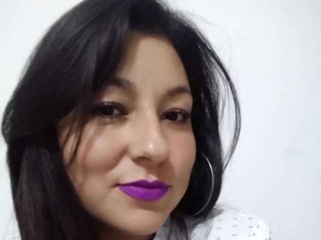 Sakoto Dika a les cheveux noirs, du rouge à lèvres violet et un léger sourire. Elle porte une chemise blanche à motifs et de grandes boucles d'oreilles. Sa peau est de couleur marron clair..