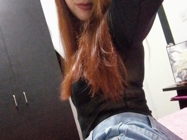 Eliza Jones, com cabelos ruivos longos, uma blusa preta e jeans azuis, exibindo suas nádegas enquanto levanta um braço. Seu rosto está parcialmente visível, com os lábios ligeiramente entreabertos..
