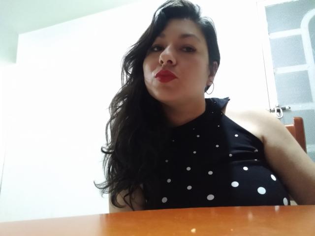 SakotoDika with dark wavy hair, red lipstick, black polka dot sleeveless top, sitting at a wooden table, looking down with a slightly pouty expression.