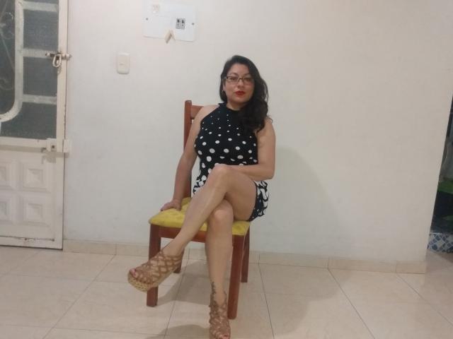 SakotoDika with black polka dot dress, red lipstick, glasses, crossed legs, yellow chair, brown sandals, white wall, door, and tile floor, sitting and looking at camera.