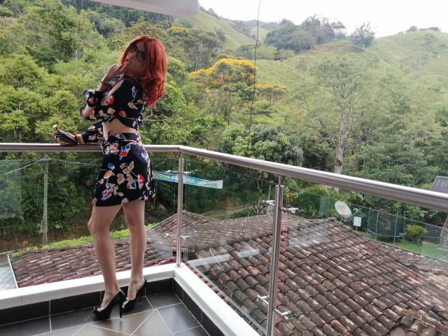 Rosariio with red hair, floral dress, black heels, and glasses, standing on a tiled balcony, holding a phone, looking down, green hills and trees in the background.
