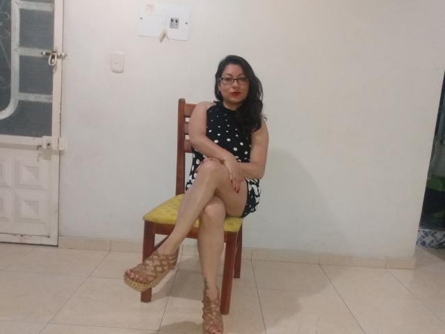 SakotoDika, with black hair, glasses, red lipstick, black polka-dot dress, and crossed legs, sits on a yellow-cushioned wooden chair, wearing brown wedge sandals, in a room.