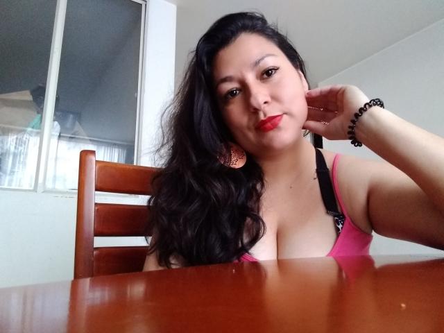 SakotoDika con cabello negro largo, blusa rosa que muestra un busto grande, lápiz labial rojo, silla de madera, pulsera de cuentas negras, con la cabeza apoyada en la mano, mirando a la cámara, una mesa en frente..