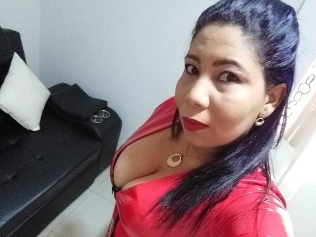 LeiizaMistress with dark blue hair, red lipstick, and red shirt showing her large boobs, wearing a gold necklace, smiling at the camera, in a room with a black couch.