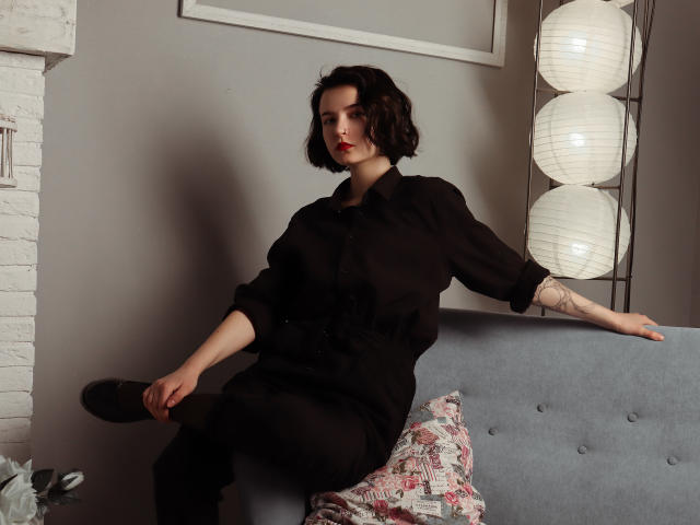 Viola, schwarzhaarig mit kurzen, schwarzen Haaren, trägt ein schwarzes Kleid und sitzt auf einem grauen Sofa. Sie hält einen schwarzen Fächer, trägt roten Lippenstift und hat ein Tattoo am rechten Unterarm. Im Hintergrund sind weiße Papierlaternen zu sehen..