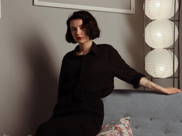 Viola, schwarzhaarig mit kurzen, schwarzen Haaren, rotem Lippenstift und einem schwarzen Hemd mit Knopfleiste, sitzt auf einem grauen Sofa, der rechte Arm ist tätowiert, der linke Arm ruht auf. Im Hintergrund sind weiße Papierlaternen zu sehen..