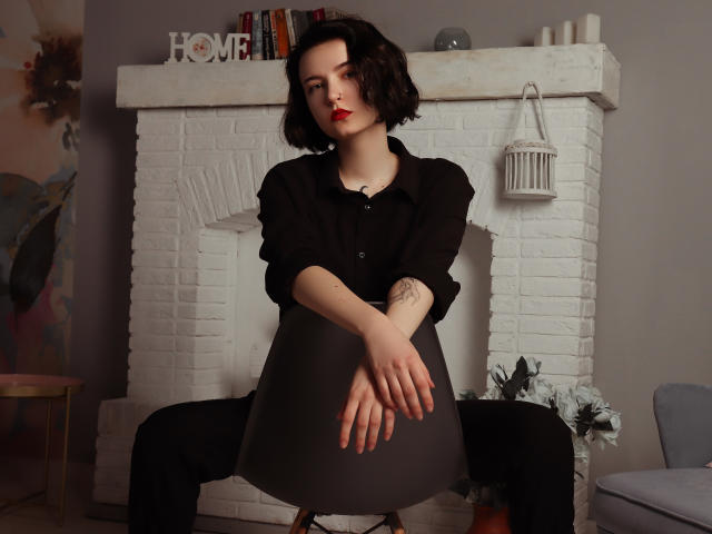Viola, cheveux noirs courts, rouge à lèvres rouge, chemise noire et jupe grise, assise devant une cheminée en briques blanches, bras croisés, tatouage sur l'avant-bras gauche, jambes écartées..