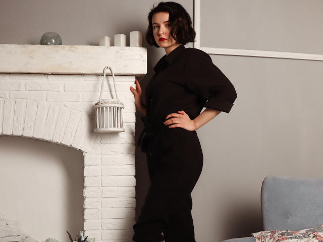 ViolaBlack está em pé, com uma mão apoiada em uma lareira de tijolos brancos, vestindo um vestido preto, batom vermelho e cabelo curto e escuro, com um candeeiro branco pendurado na lareira..