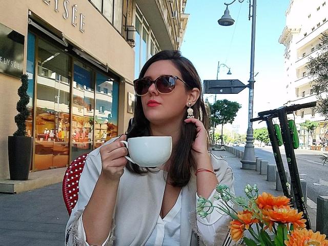 ChristineRayssa, con cabello castaño, usando gafas de sol, sosteniendo una taza blanca, vestida con una camisa blanca, sentada cerca de flores naranjas, en una calle soleada, tocándose el cabello, con una pulsera roja..