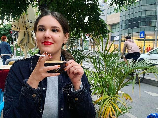 ChristineRayssa met rode lippenstift, die een kop koffie vasthoudt, gekleed in een denimjas, staat buiten, sigaret rokend, groene boom op de achtergrond, stadsstraat, witte auto, blauw bord..