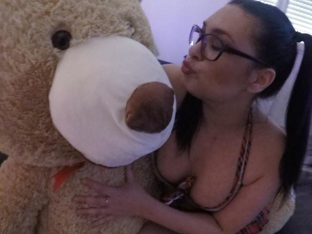 SelinaKylla, con gli occhiali, sta baciando il naso di un enorme orsacchiotto beige, con il décolleté visibile in un top a halter con motivo, e le mani sul muso peloso dell'orsacchiotto..